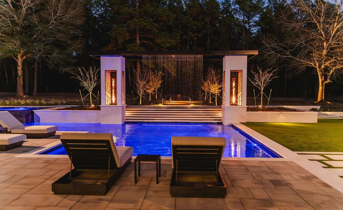 Backyard pool at night with a sheer waterfall between two lit pillars and lounge chairs on the patio